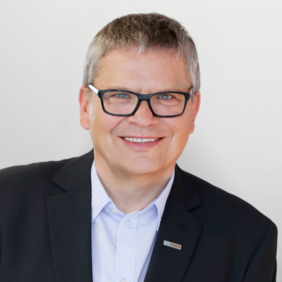 A smiling middle-aged man with short gray hair and black glasses is wearing a dark blazer over a light blue shirt. He appears friendly and approachable, with a neutral light background. A small name tag is visible on his blazer.