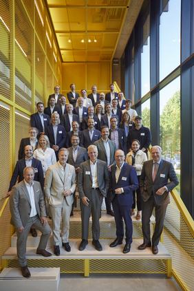 Eine Gruppe von etwa 40 Personen steht auf einer Treppe in einem modernen Gebäude mit großen Fenstern. Sie tragen formelle Kleidung und lächeln für das Foto. Die Wände sind gelb und der Raum ist hell erleuchtet.