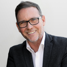 A smiling man with short, dark hair and glasses is wearing a dark blazer over a white shirt. He is leaning slightly to one side, with a neutral background, conveying a friendly and approachable demeanor.