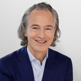 A man with medium-length, wavy gray hair smiles warmly. He wears a dark blue blazer over a light blue shirt. The background is light and simple, emphasizing his friendly expression.