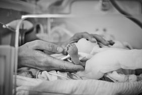 Ein Hand greift zärtlich nach einem kleinen Fuß eines Neugeborenen, das in eine Decke gewickelt ist. Die Szenerie vermittelt Geborgenheit und Fürsorge. Es ist eine emotionale Szene, die die Verbindung zwischen dem Erwachsenen und dem Baby zeigt.