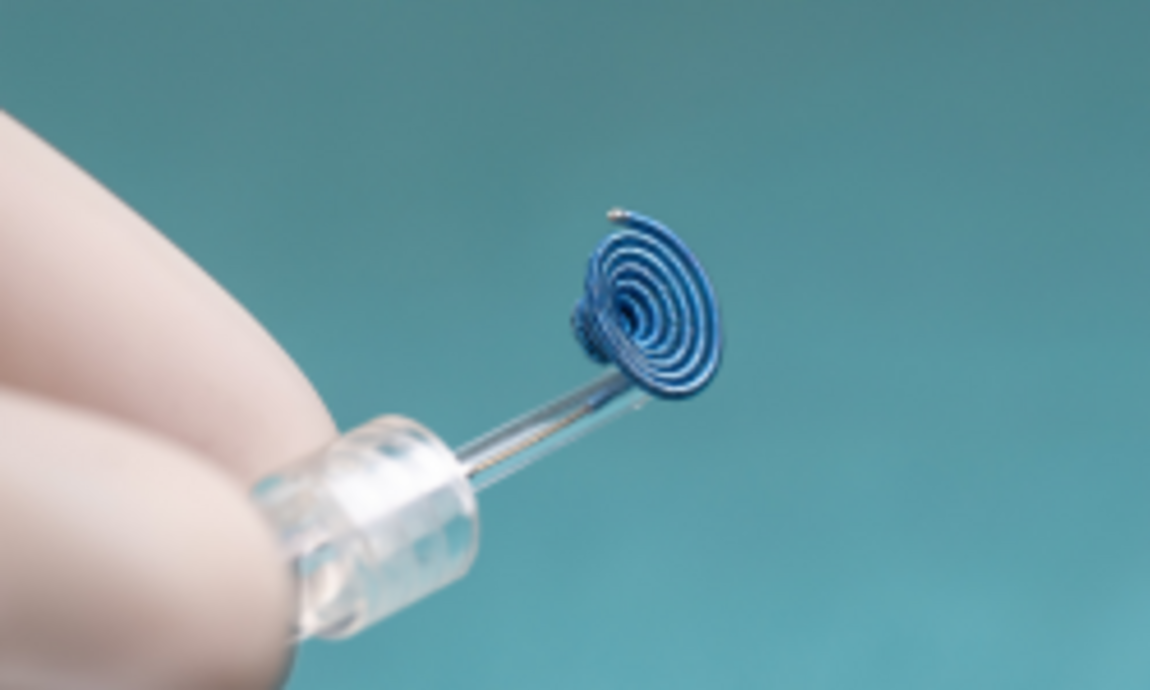 Eine Hand hält einen dünnen, transparenten Behälter, aus dem eine spiralförmige Struktur aus blauen, gedrehten Drahte heraussieht. Der Hintergrund ist einfarbig und blau. Die Spirale scheint klein und präzise verarbeitet zu sein.