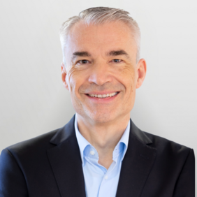 Ein lächelnder Mann mit grauen Haaren trägt einen schwarzen Anzug und ein hellblaues Hemd. Der Hintergrund ist neutral und hell.
