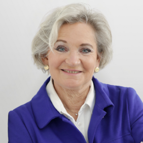 A smiling older woman with short gray hair is wearing a purple jacket over a white shirt. She has pearl stud earrings and appears confident, with her arms crossed in front of her. The background is light and neutral.