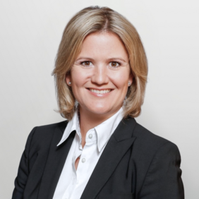 Portrait einer Frau mit blonden, schulterlangen Haaren, die in einem schwarzen Blazer und weißem Hemd gekleidet ist. Sie lächelt freundlich und schaut direkt in die Kamera. Der Hintergrund ist hell und schlicht.