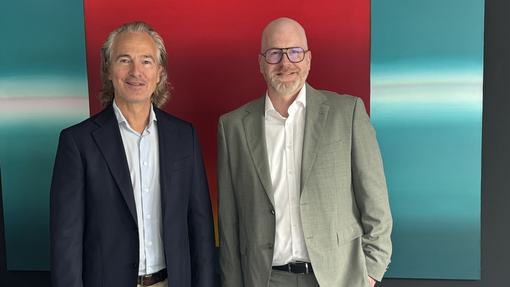 Zwei Männer stehen nebeneinander vor einem farbigen Hintergrund mit einem großen roten und türkisfarbenen Bereich. Der linke Mann hat längere, lockige Haare und trägt eine dunkle Jacke. Der rechte Mann ist glatzköpfig und trägt einen grauen Anzug. Beide lächeln freundlich.