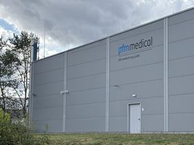Das Bild zeigt die Außenansicht eines modernen, grauen Unternehmensgebäudes mit dem Schriftzug "pfm medical" an der Wand. Vor dem Gebäude ist eine grüne Wiese mit Büschen zu sehen. Der Himmel ist bewölkt.