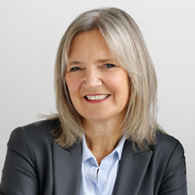 A smiling woman with shoulder-length gray hair is wearing a black blazer over a light blue shirt. She is facing the camera against a neutral background, conveying a friendly and professional demeanor.