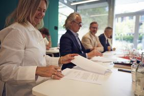 Eine Gruppe von fünf Personen sitzt an einem Tisch. Eine Frau in einem weißen Hemd blättert durch Dokumente. Die anderen vier Männer sind in formeller Kleidung gekleidet und hören aufmerksam zu. Im Hintergrund sind Fenster und Pflanzen sichtbar.