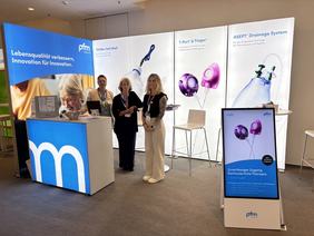 Messestand mit drei Personen, die freundlich zusammenstehen. Im Hintergrund sind großflächige Banner mit Informationen über medizinische Produkte sichtbar. Ein Bildschirm zeigt zusätzlich Informationen zu einem Drainagesystem. Der Stand vermittelt einen professionellen Eindruck im Bereich Gesundheitsinnovation.
