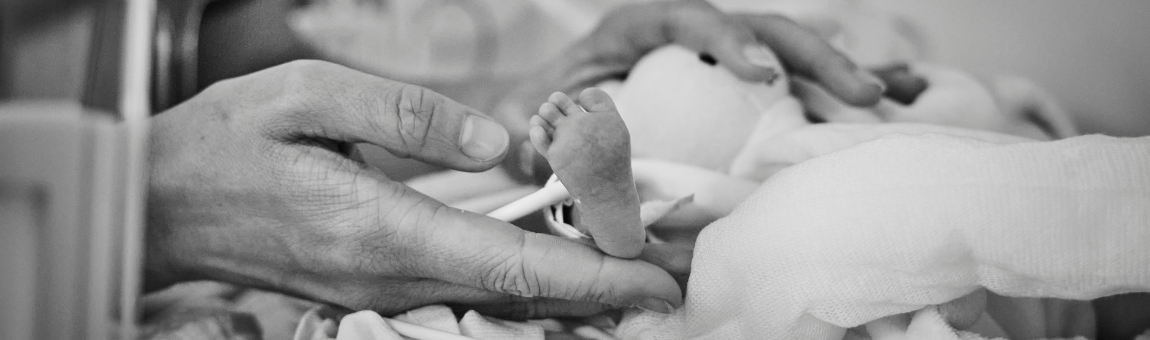 Ein neugeborenes Babyfußchen wird zärtlich von einer Hand gehalten. Es zeigt die sanfte Verbindung zwischen einem Elternteil und dem Kind, umgeben von Binden und Decken in einem medizinischen Umfeld. Die Szene vermittelt Gefühle von Sorge und Zuneigung.
