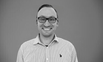 Ein lächelnder Mann mit Brille trägt ein gestreiftes Hemd. Der Hintergrund ist neutral und einfarbig. Der Mann steht mittig im Bild und strahlt Freundlichkeit und Offenheit aus.