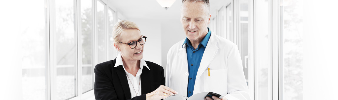 A woman in business attire and a man in a lab coat are discussing information from a document while standing in a well-lit hallway. The woman points to the document, and they both appear engaged in conversation.